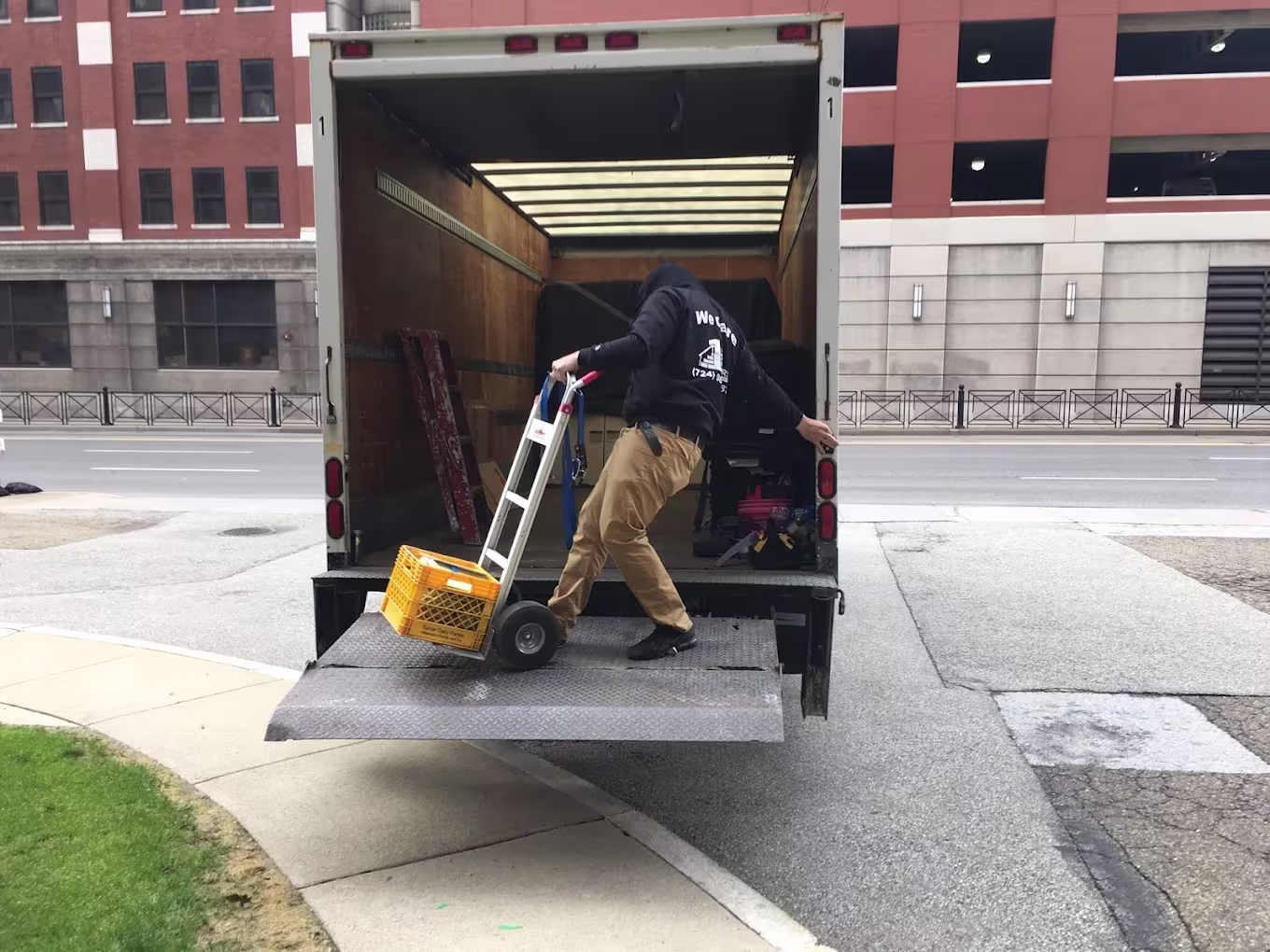 Mover using a hand truck to roll items up the lift gate into a 1st Step truck on a city street